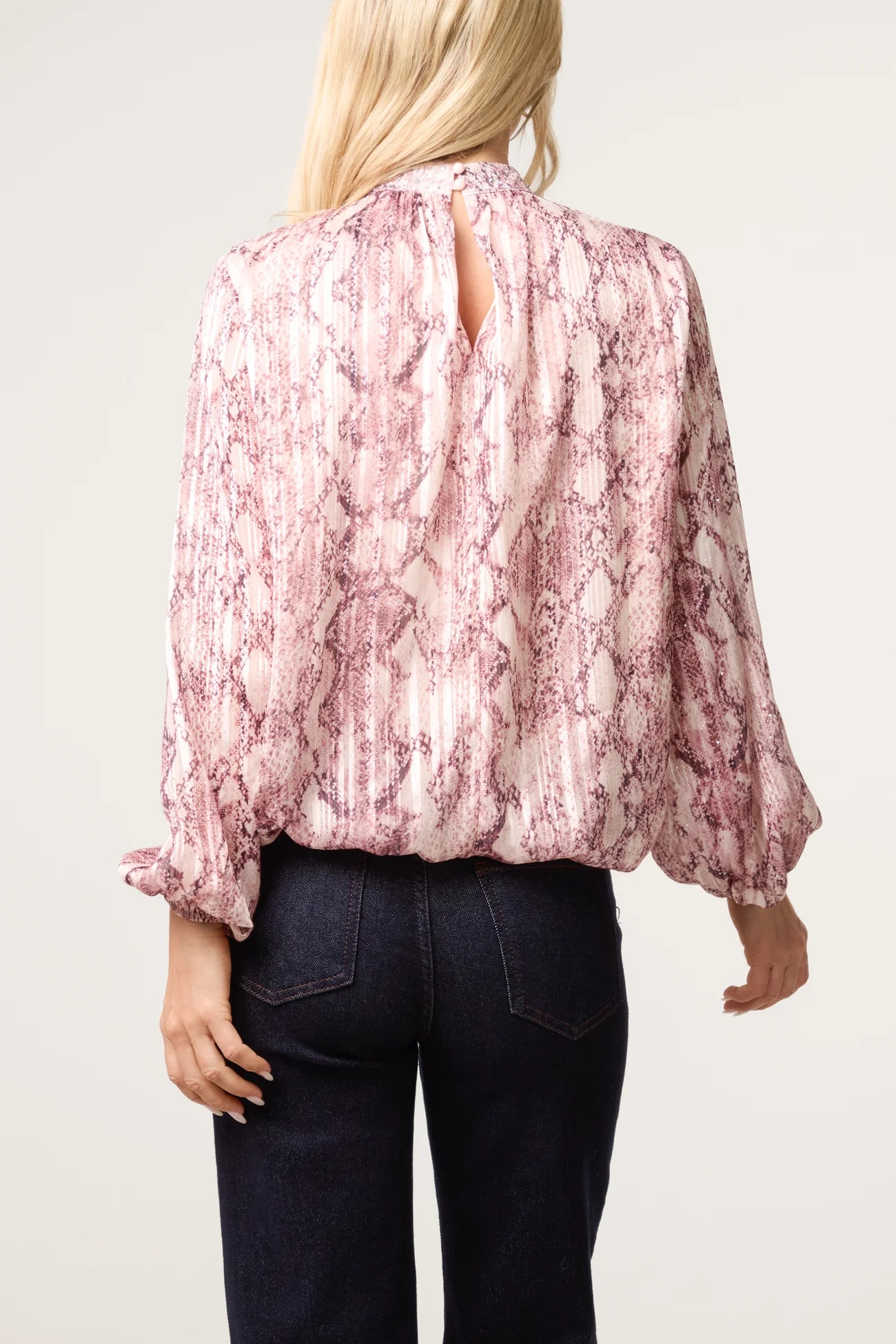 A woman shown from the back wearing a pink and white snakeskin print blouse with long balloon sleeves and a keyhole back, paired with dark blue jeans, against a light gray studio background.