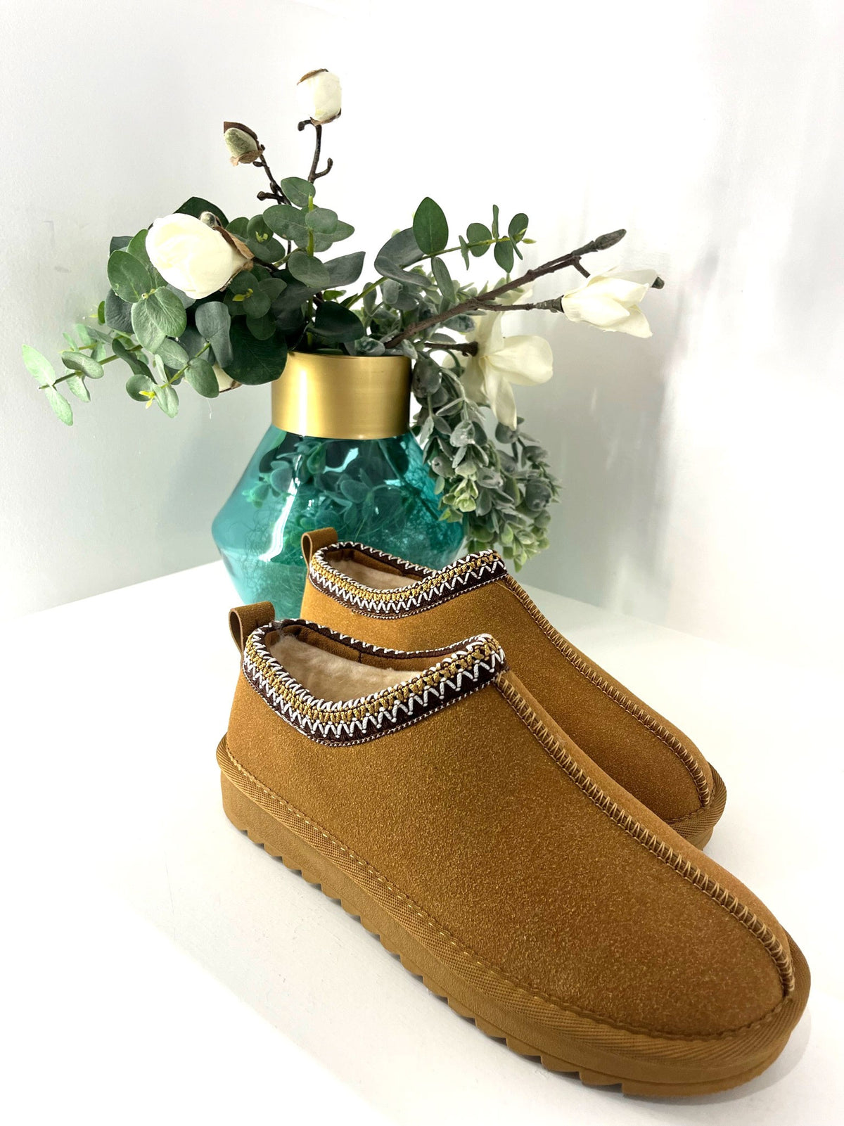 A pair of tan suede slip-on slippers with a decorative black, white, and gold patterned trim around the collar, displayed on a white surface with a green glass vase of flowers in the background.